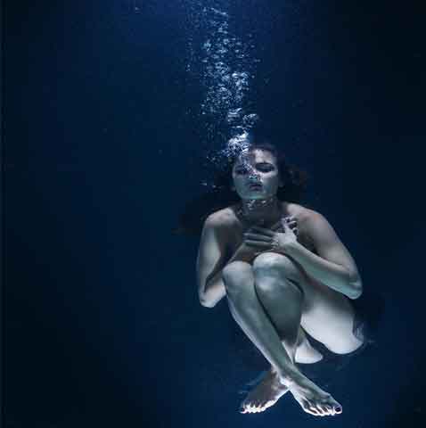 a nude woman in a fetal position beneath blue water who is sinking deeper
