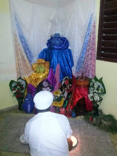 Alo Ocan Roberto Gonzalez Sousa in front of his Orish Altar. Image of Alo Ocan Roberto Gonzalez sitting with his back facing us, in front of him is a religious altar with a which decorative dove.